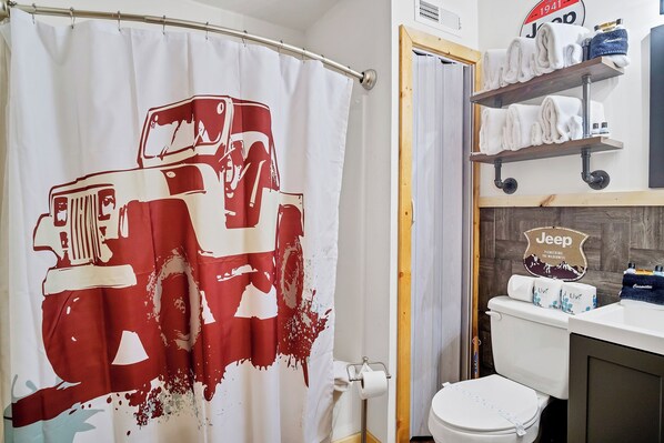 A bathroom with a shower curtain featuring a red and white vehicle design, a toilet, and a shelf with towels and toiletries.