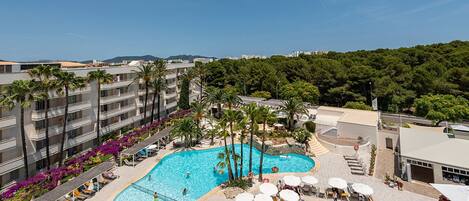 A rooftop pool area with lounge chairs and umbrellas, surrounded by palm trees and a view of the city.