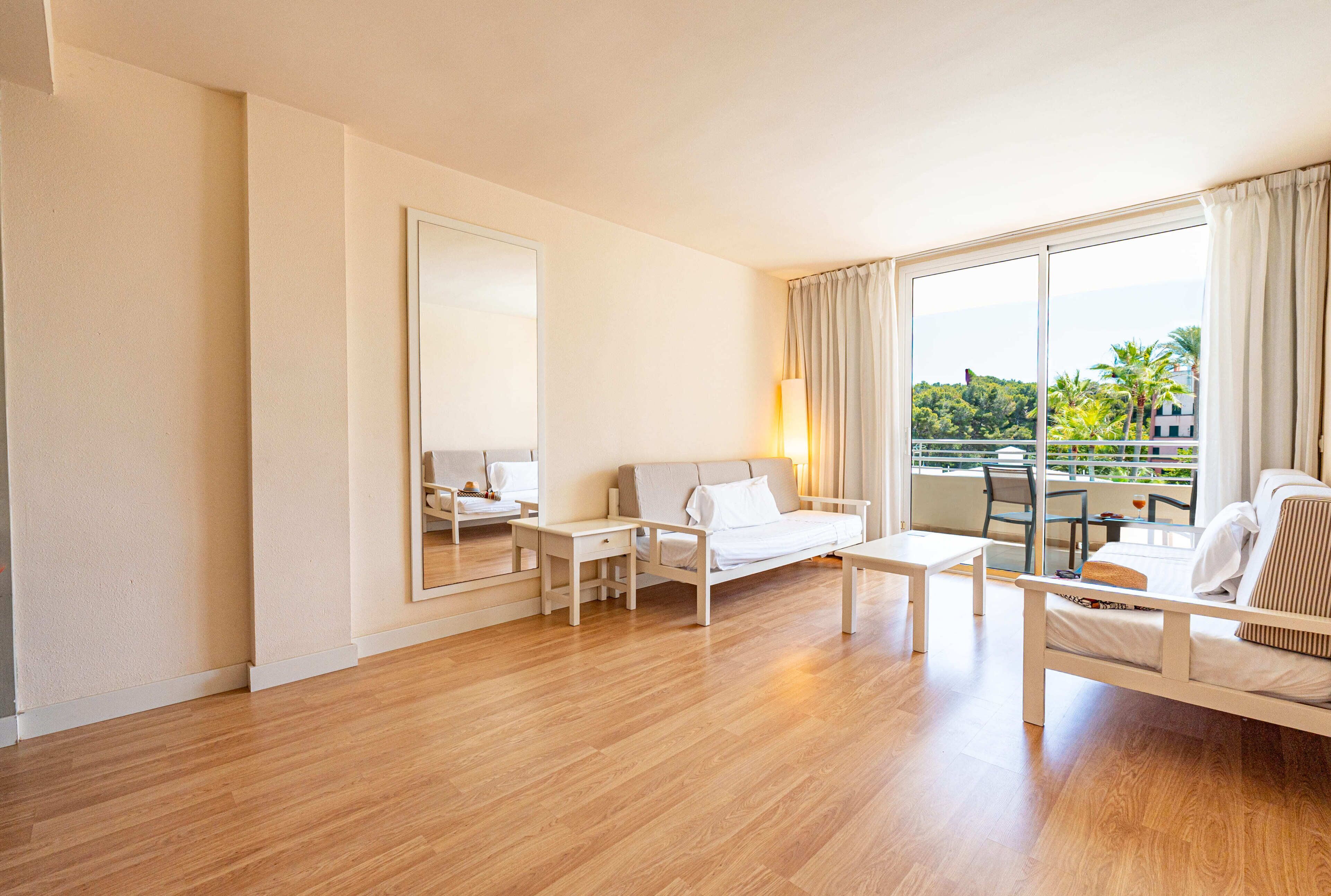 A hotel room with a wooden floor, a bed, a sofa, a table, a chair, a large mirror, and a balcony with a view of greenery.