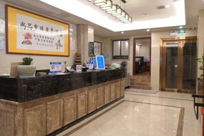 Un espace d’accueil d’hôtel comprenant un comptoir de réception, des œuvres d’art encadrées, un écran numérique et un escalier menant à l’entrée.
