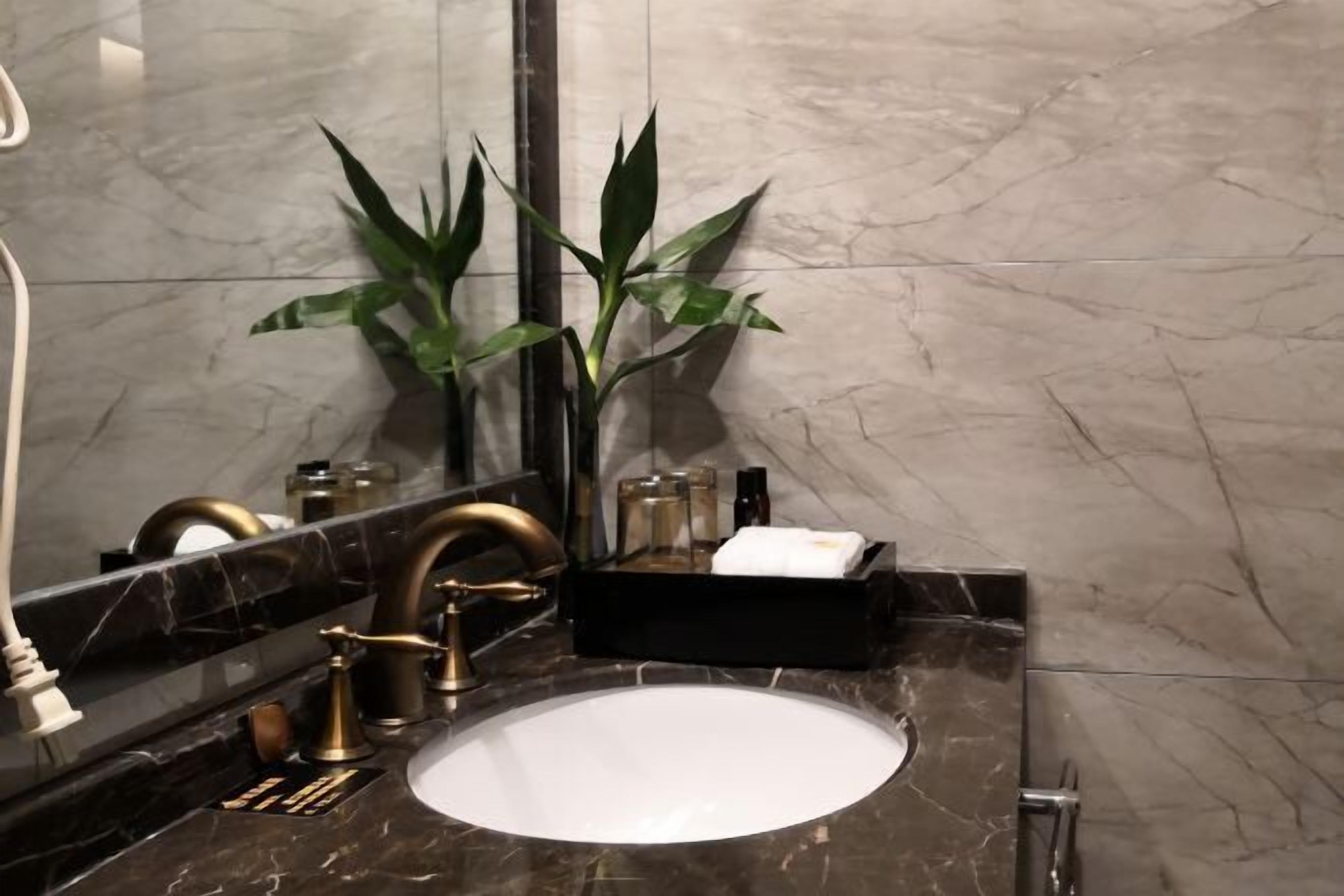 Ein Badezimmer mit Marmorwaschtisch, goldenem Wasserhahn und einem Spiegel. Auf der Ablage steht ein Topfpflanze und eine schwarze Seifenschale.