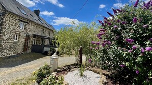 Une maison en pierre avec une allée gravillonnée, un jardin où poussent différentes espèces de plantes et un ciel bleu azur.
