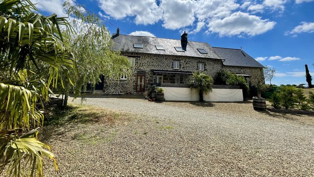 Une maison en pierre avec une allée de gravier, un petit jardin et un ciel bleu dégagé.