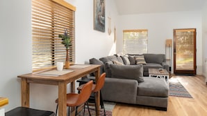 En stue med en grå sofa, træpersienner, et spisebord med stole og et billede på væggen.