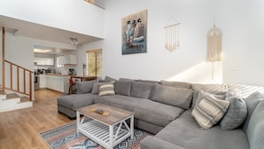 En moderne stue med en grå hjørnesofa, et træbord og et mønstret tæppe. Et billede og en macramé-vægophængning pryder væggen.
