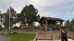 A stone pathway leading to a wooden gazebo with a flagpole, surrounded by a well-maintained lawn and trees. - Gorgeous Log Cabin estate with hot tub, views, privacy and sauna ! (Durango)