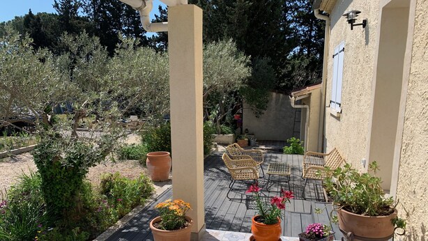 Een terras buiten met een betegelde vloer, planten in potten, rieten meubels en een pergola.