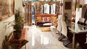 A living room with a sofa, a dining table, and a glass coffee table. There are curtains, a chandelier, and a potted plant.