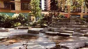 A stone fountain with water cascading down steps, surrounded by greenery and urban buildings.