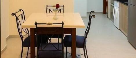 A dining area with a table and chairs, a painting on the wall, and a kitchen in the background.
