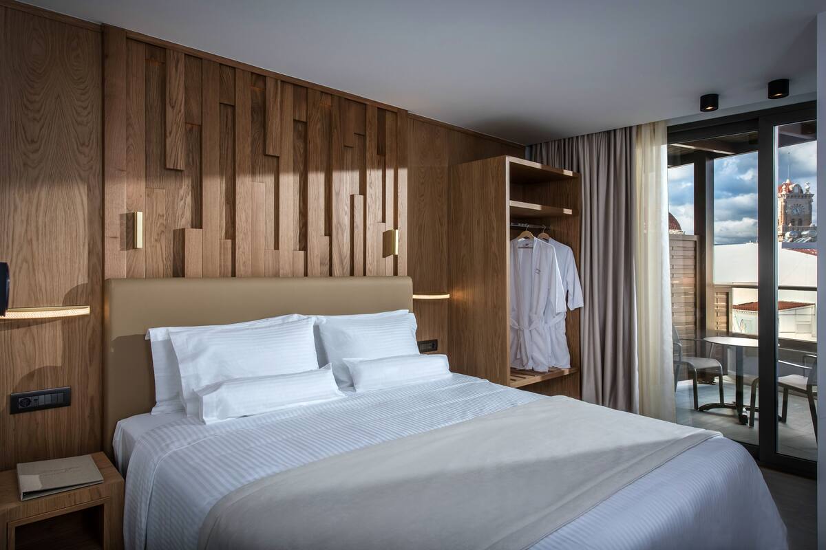 Een moderne hotelkamer met een groot bed, een wandpaneel van hout, een kledingkast en een balkon met uitzicht op een klokkentoren.
