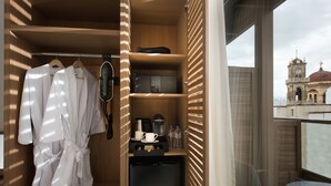 Hotelkamer met een kledingkast met badjassen, een haardroger en een kleine koelkast. Er is een klein tafeltje en een stoel, en uitzicht op een gebouw met een klokkentoren.
