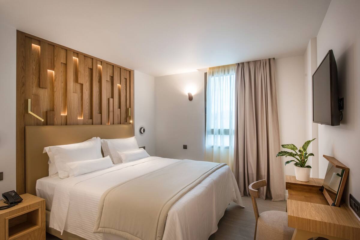 Een moderne hotelkamer met een houten hoofdeinde, een bed met witte lakens, een nachtkastje met een telefoon, een bureau met een plant en een aan de muur bevestigde televisie.