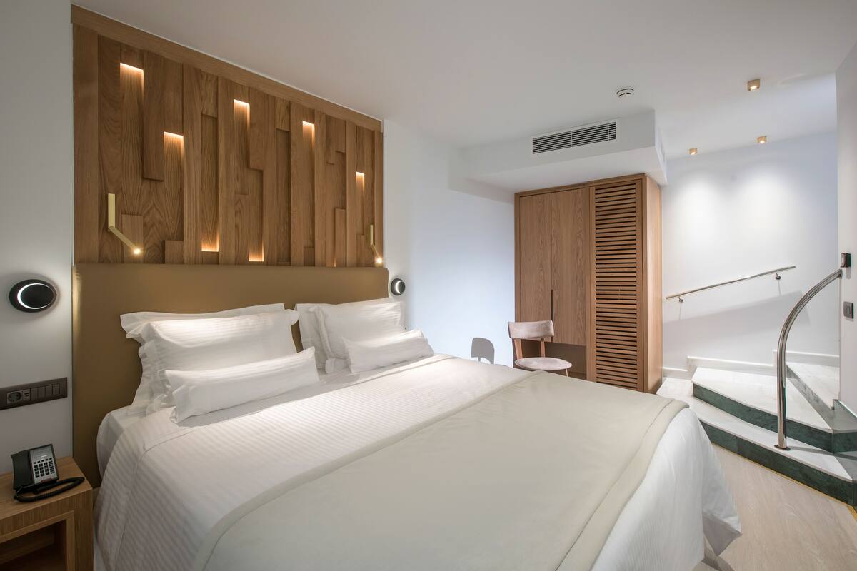 Een moderne hotelkamer met een groot bed, een houten hoofdbord met verlichting, een klein nachtkastje met een telefoon en een trap met een leuning.