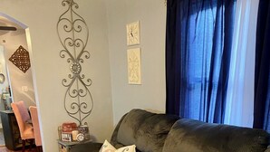 Un salon avec un canapé gris foncé, un plaid rose et un jeté en tricot. Le sol est en bois et, au fond, on aperçoit un rideau bleu.