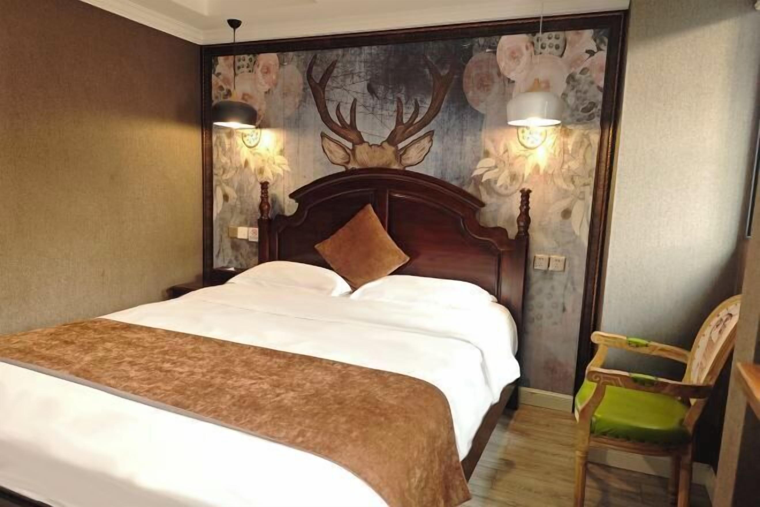 Ett hotellrum med en stor säng, ett träfärgat sänggavel, två vägglampor, en blommig tapet och en grön stol.