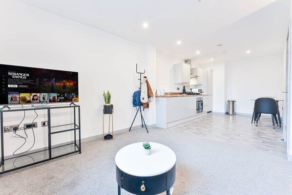Un salon moderne avec un téléviseur à écran plat, une table basse ronde et un coin cuisine à l’arrière-plan.