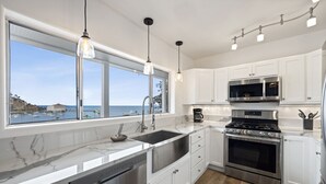 A modern kitchen with white cabinets, stainless steel appliances, a large window with a view of the sea, and pendant lighting.