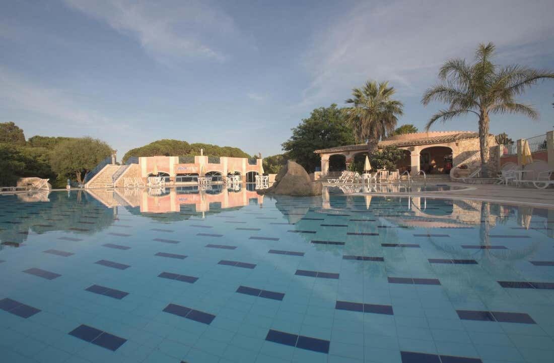 A large outdoor swimming pool with a tiled floor, surrounded by palm trees and a stone building with a covered area.