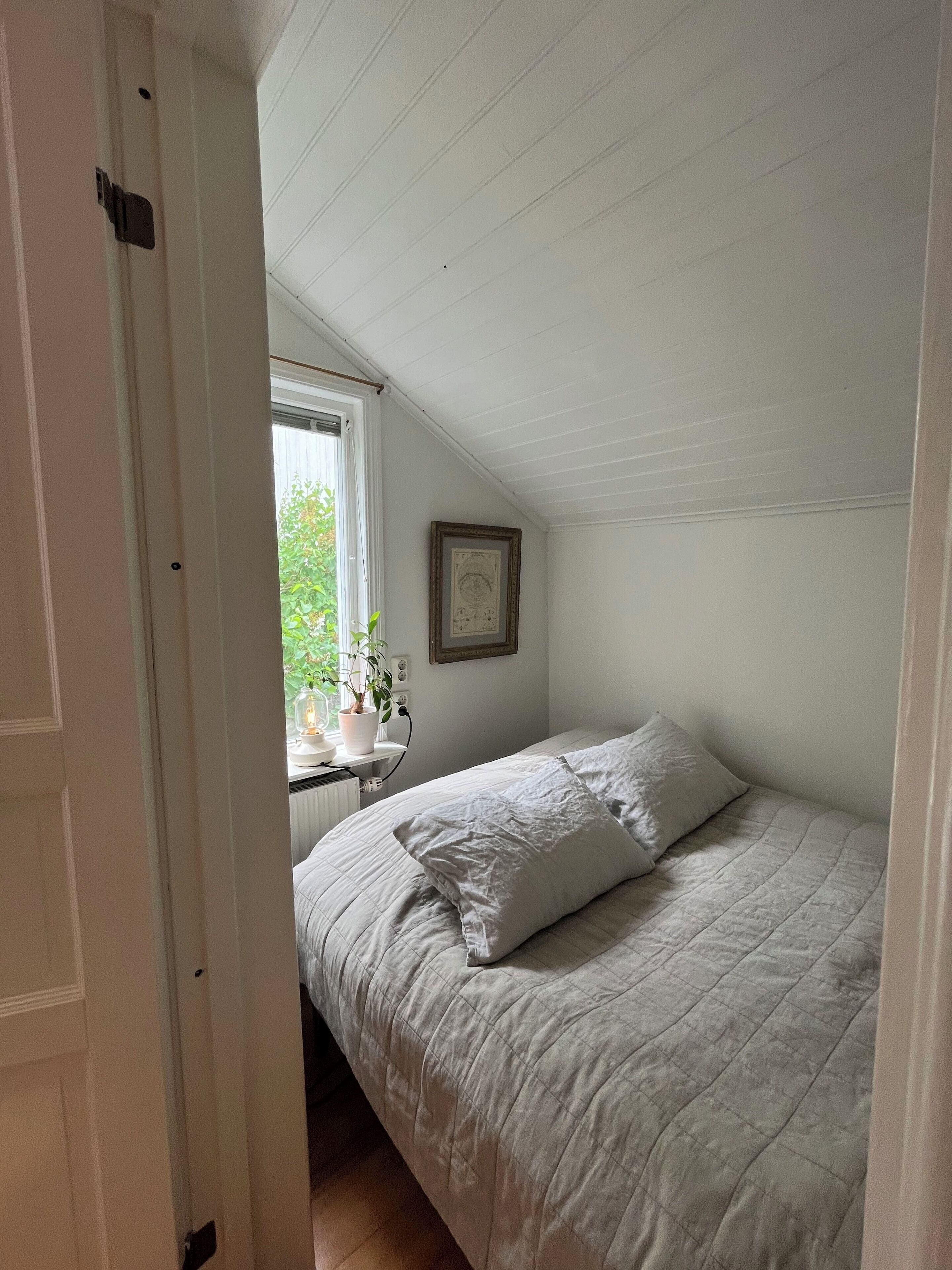 Een slaapkamer met een bed, een raam met uitzicht op groen, een lijst met een afbeelding aan de muur, en een radiator.
