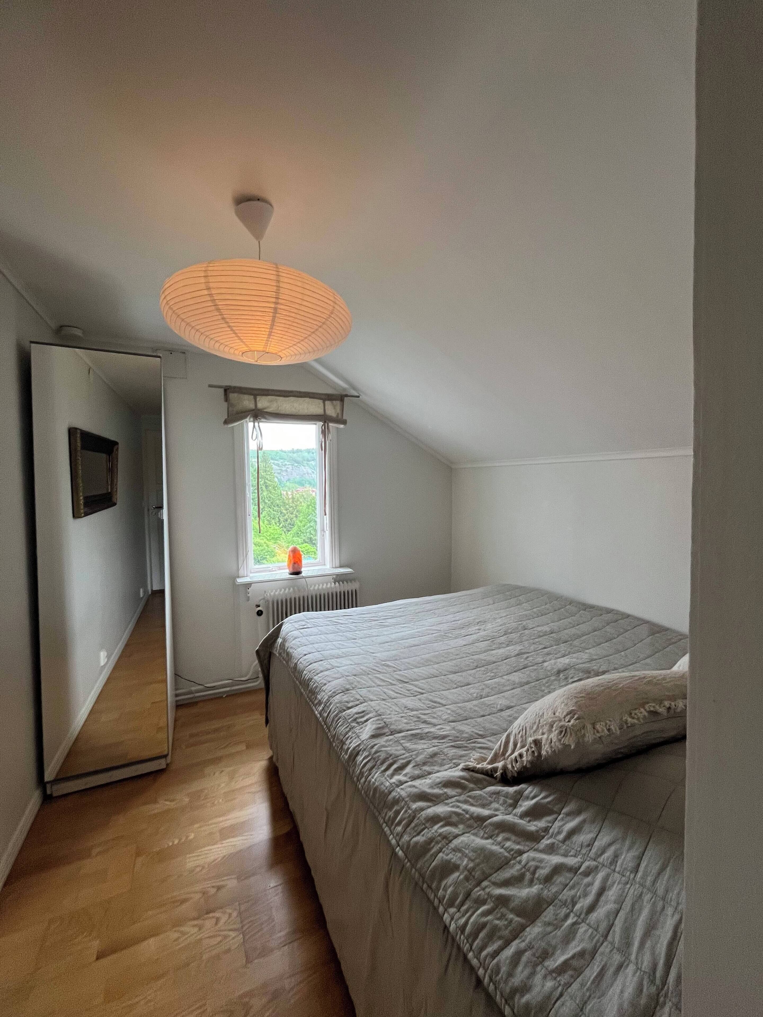 A bedroom with a bed, a window with a view, a radiator, a mirror, and a ceiling light fixture.