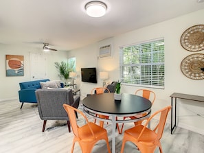 Ein modernes Wohnzimmer mit einem runden Esstisch, orangefarbenen Stühlen, einem blauen Sofa und einem grauen Sessel. An der Wand ist ein Fernseher angebracht, ein großes Fenster mit Jalousien und ein Deckenventilator.