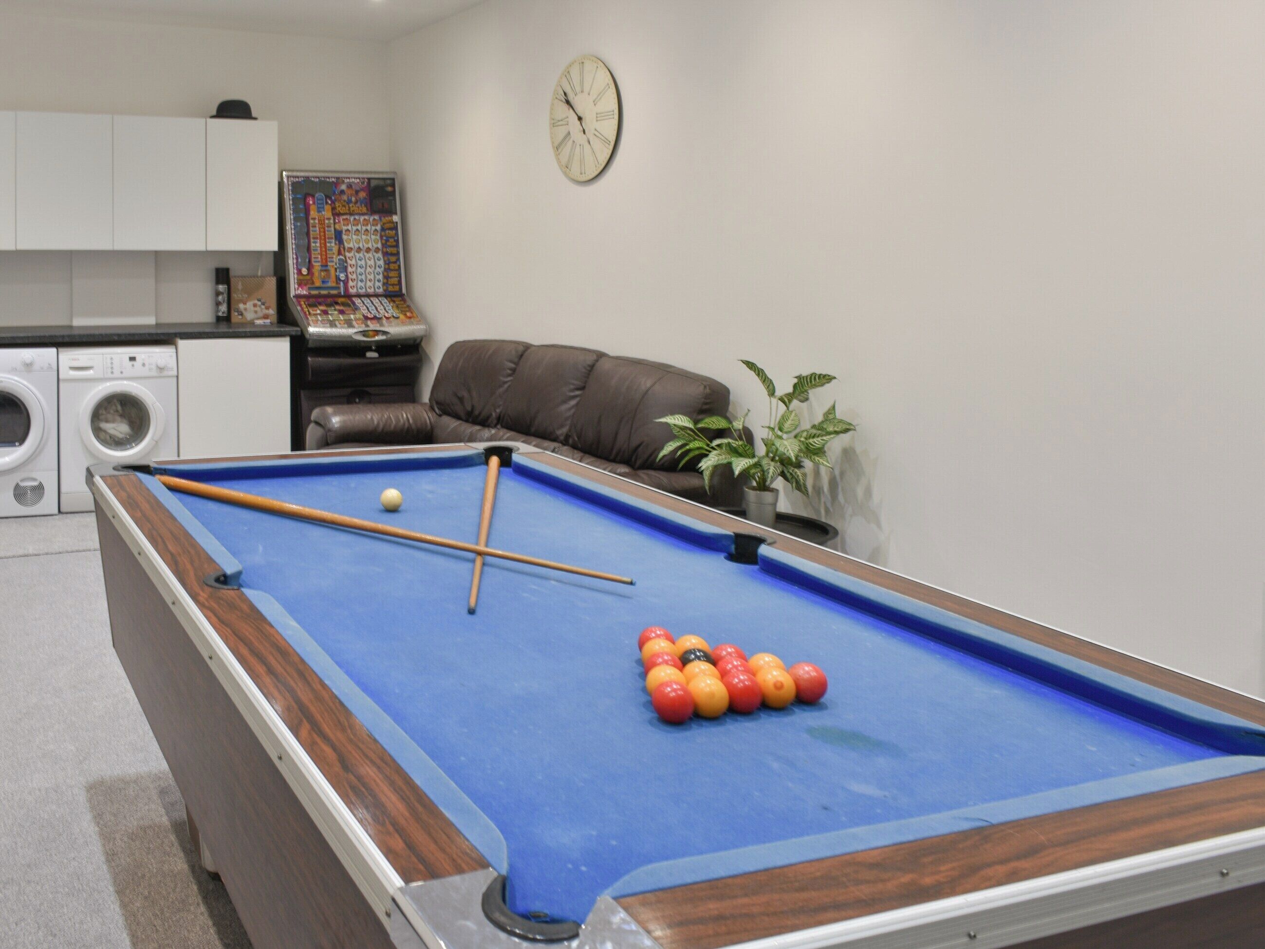 A pool table with balls, a clock, a sofa, and a potted plant in a room with a washing machine and a game console.