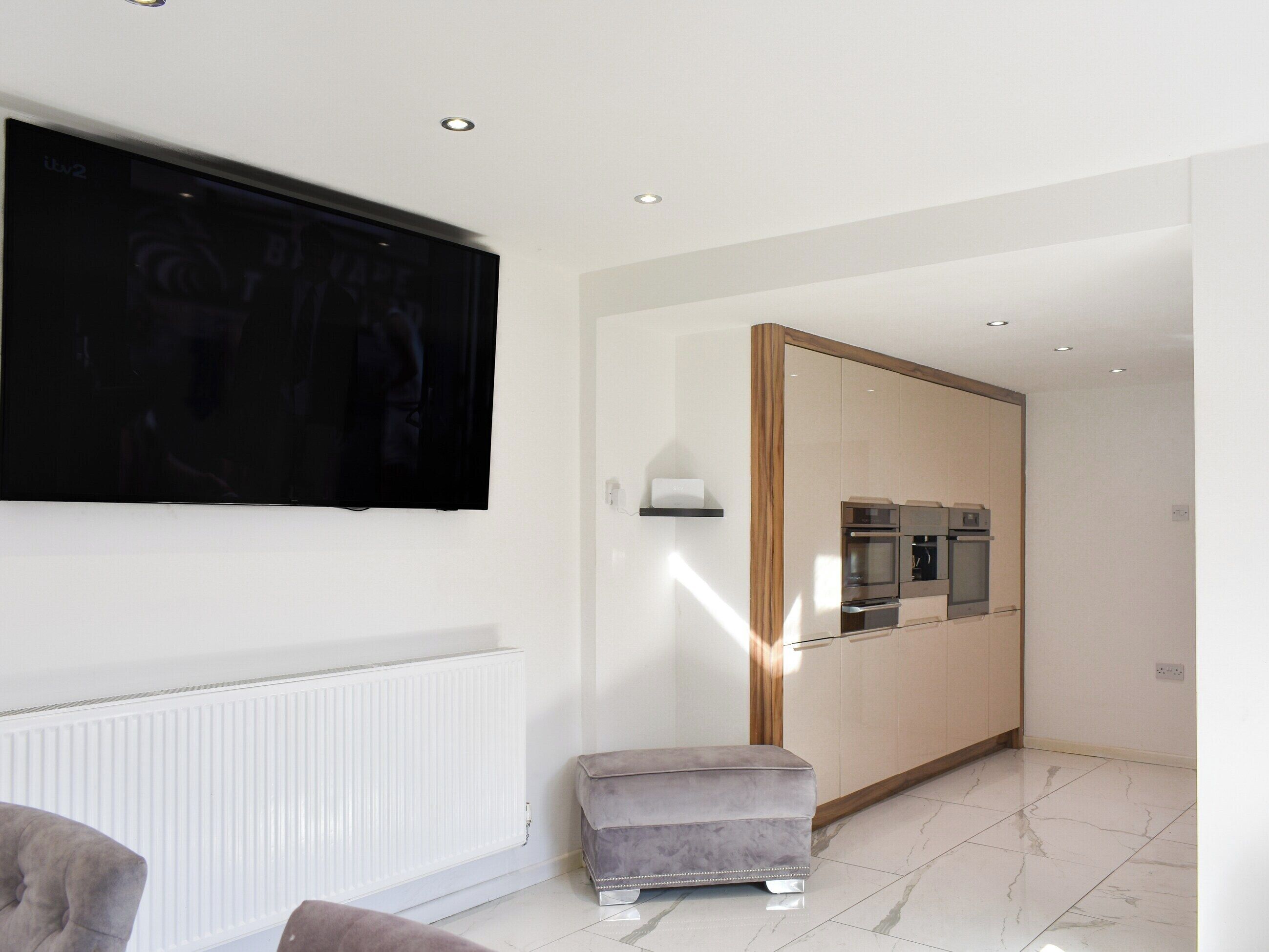 Una sala moderna con un televisor de pantalla plana, un radiador, un pequeño otomaní y una cocina con electrodomésticos integrados.