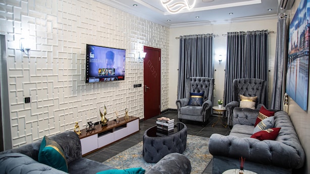 Una sala de estar moderna con un televisor de pantalla plana, sofás grises, un sillón capitonado y una pared con estampado.