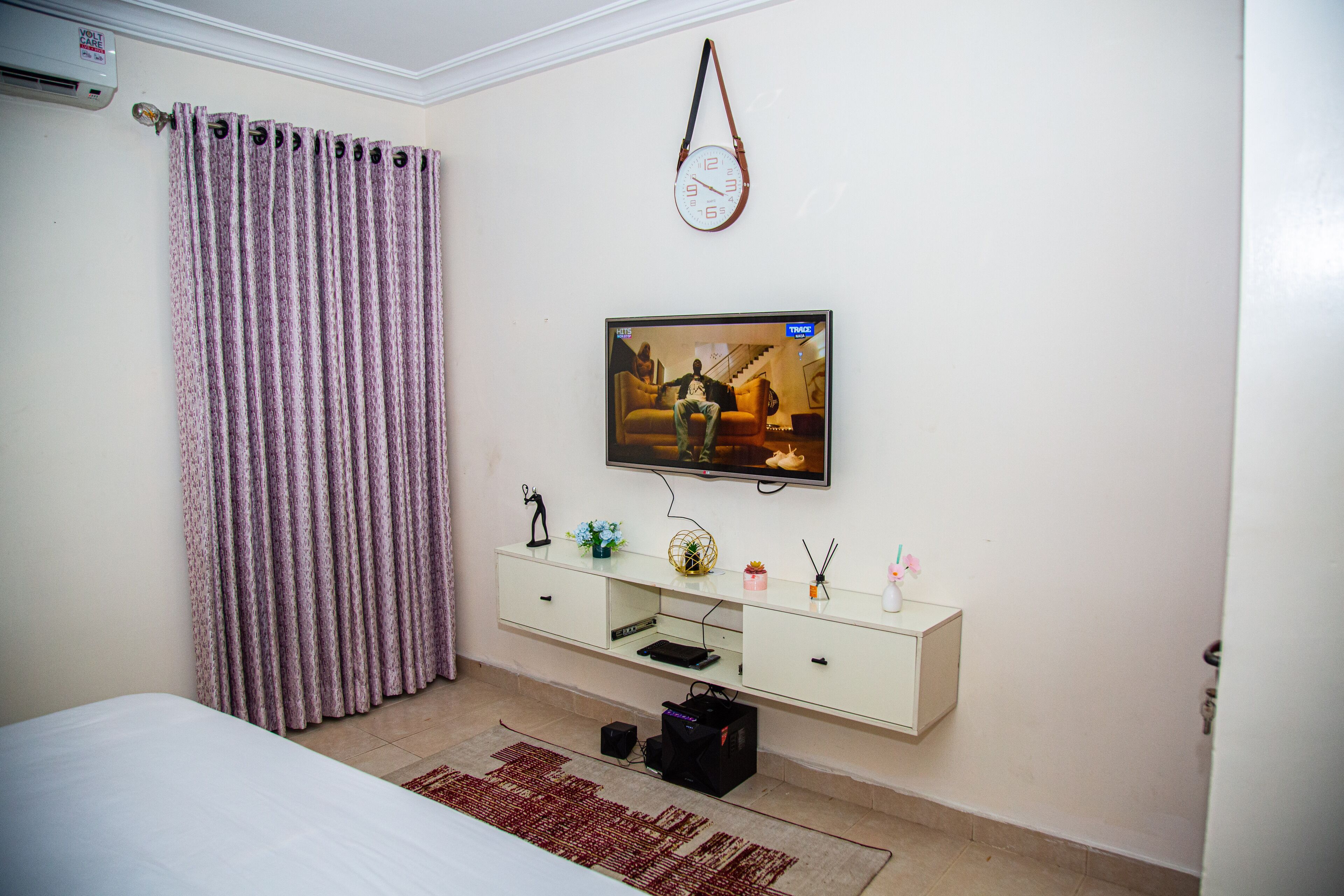 A bedroom with a bed, a TV mounted on the wall, a white dresser, and a curtain.