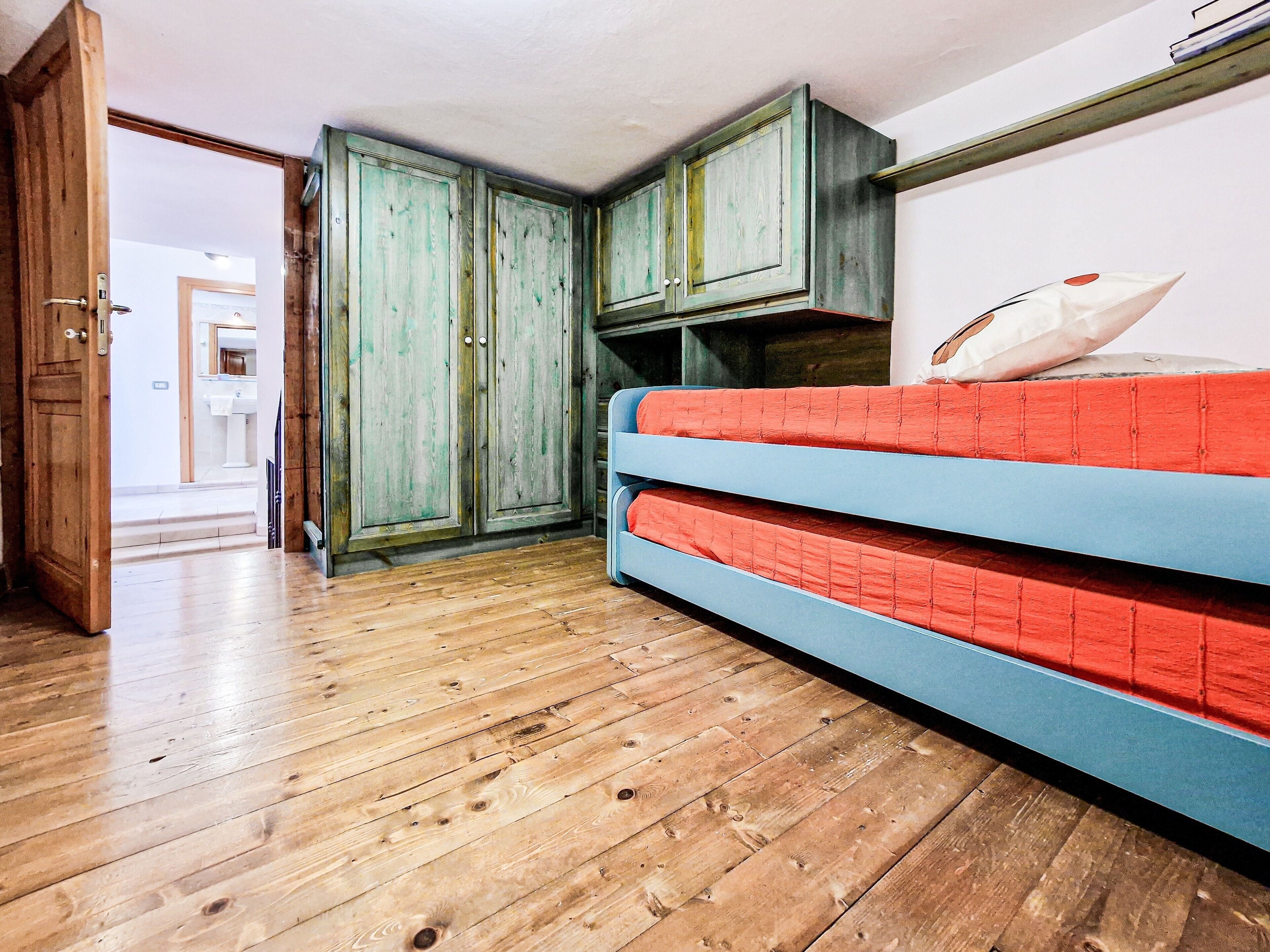 A room with wooden flooring, a bed with red and blue bedding, and green-painted cabinets.