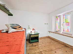 Ein Zimmer mit einem Bett, einem Nachttisch mit Lampe, einem Fenster mit bunten Vorhängen und Holzboden.