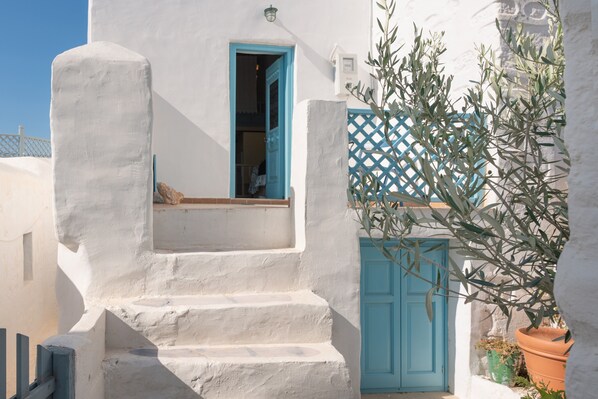 Un bâtiment blanc avec une porte bleue et une fenêtre bleue, un escalier menant à un balcon, et un olivier en pot.