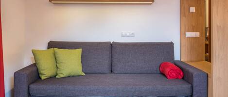Um sofá cinzento com duas almofadas, um cobertor vermelho e uma luminária fixada na parede.
