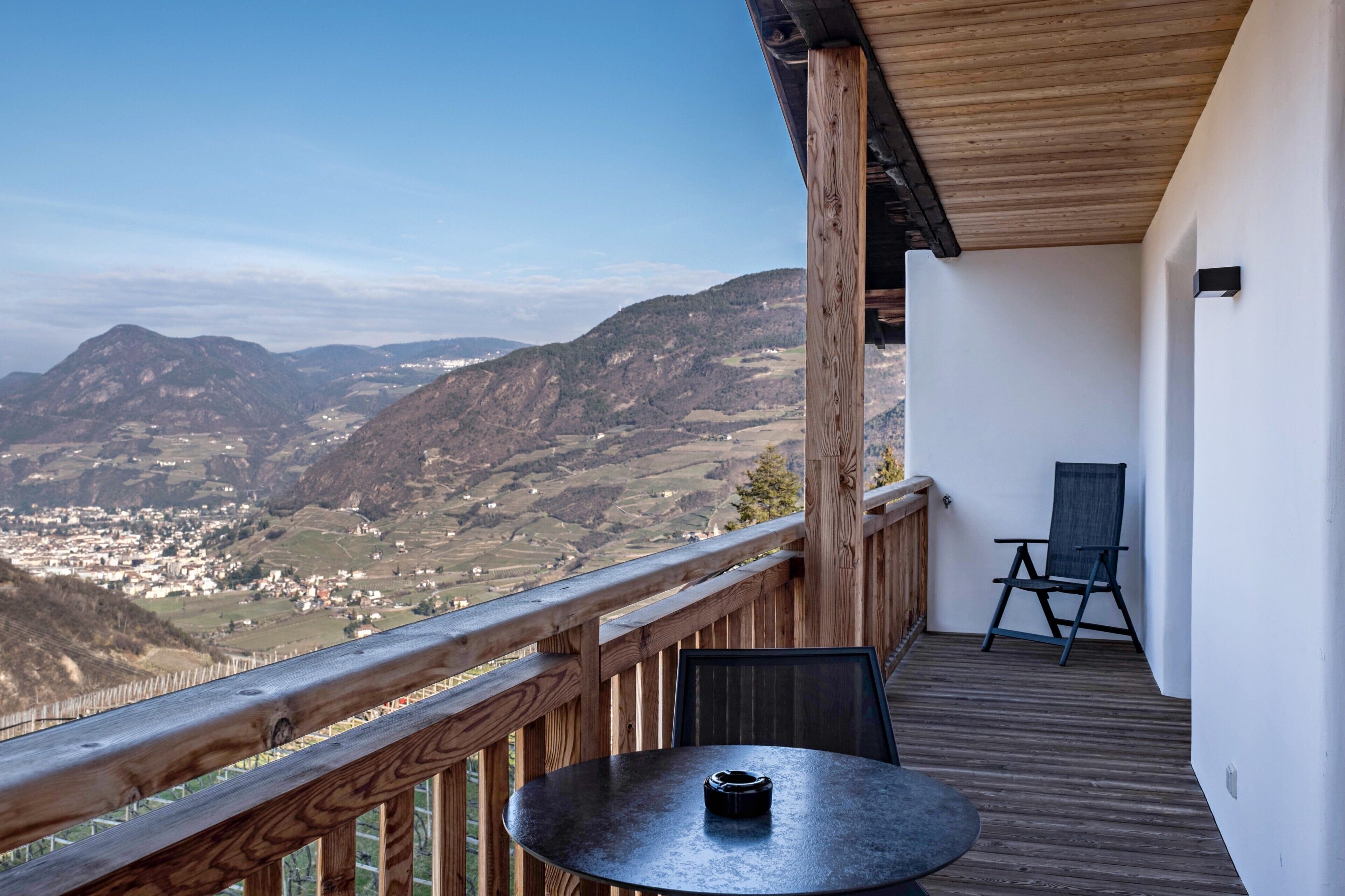 Un balcón con barandilla de madera, una mesa y una silla, con vistas a un paisaje montañoso.