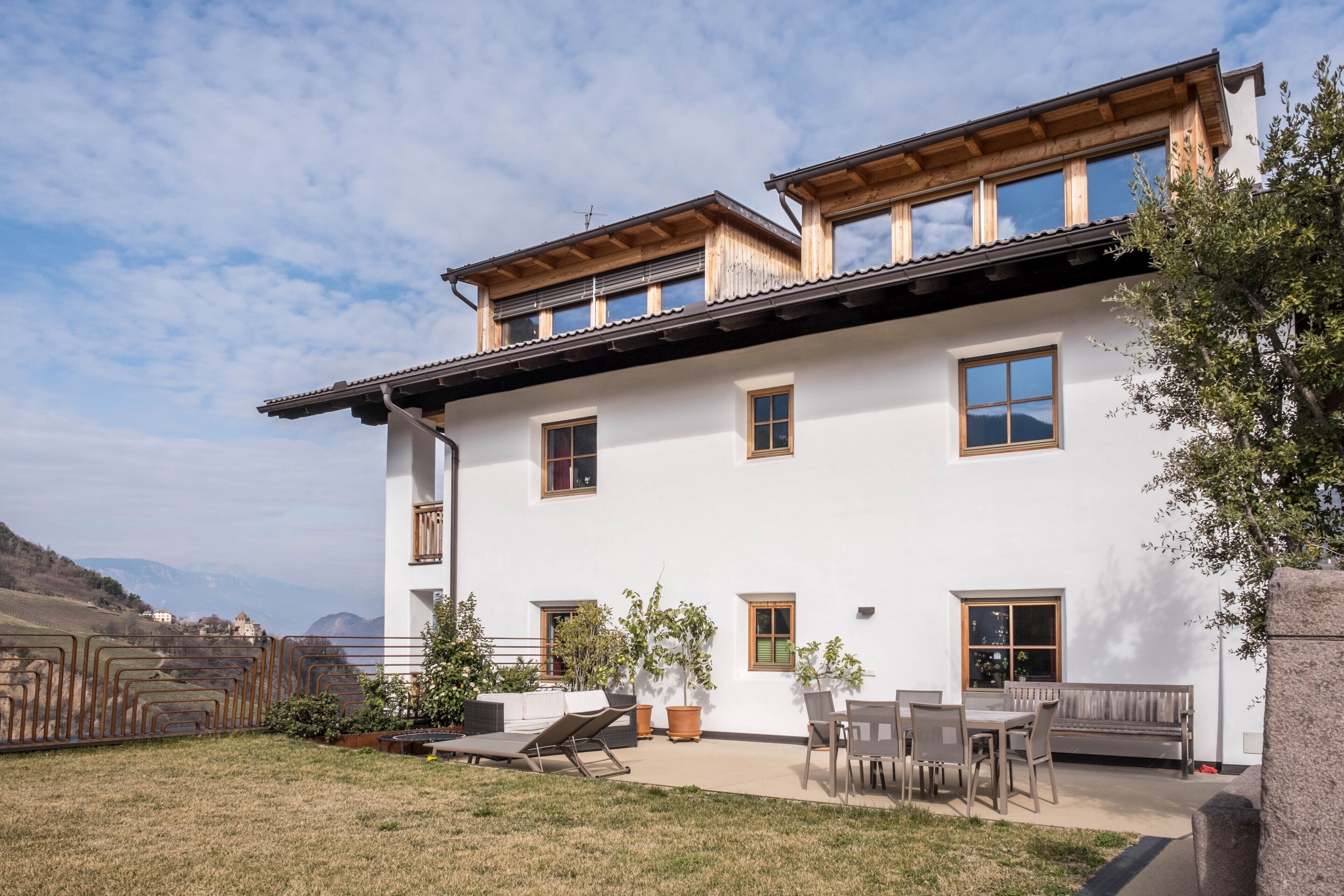 Un edificio de dos pisos con un balcón, un patio con mobiliario exterior y vistas del paisaje circundante.