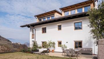 Un edificio de dos pisos con un balcón, un patio con mobiliario exterior y vistas del paisaje circundante.