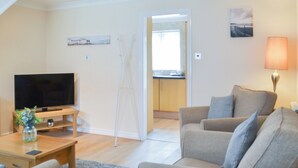 Ein Wohnzimmer mit Fernseher, Sofa und Lampe. Durch eine offene Tür ist eine Küche erkennbar.