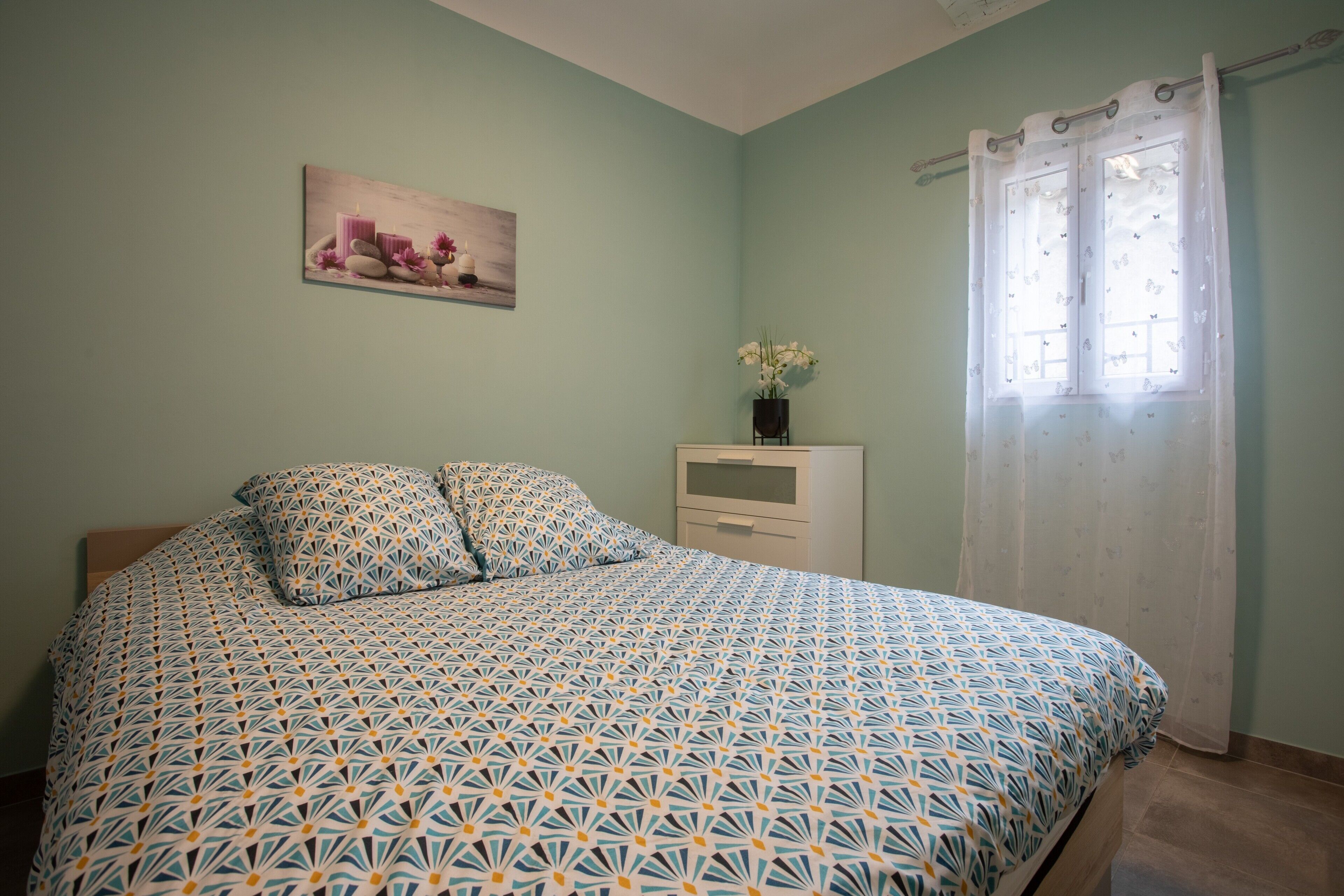 A bedroom with a patterned bedspread, a nightstand with a vase of flowers, a painting on the wall, and a window with a curtain.