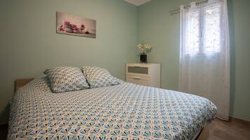Una camera da letto con una coperta dal disegno decorativo, un comodino con un vaso di fiori, un quadro al muro e una finestra con tenda.
