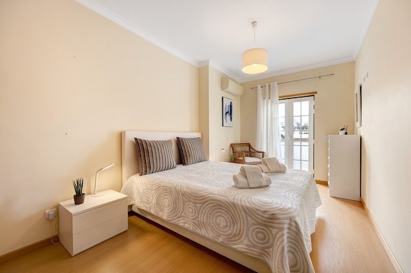 A bedroom with a bed, bedside table, lamp, and a view of the outside.