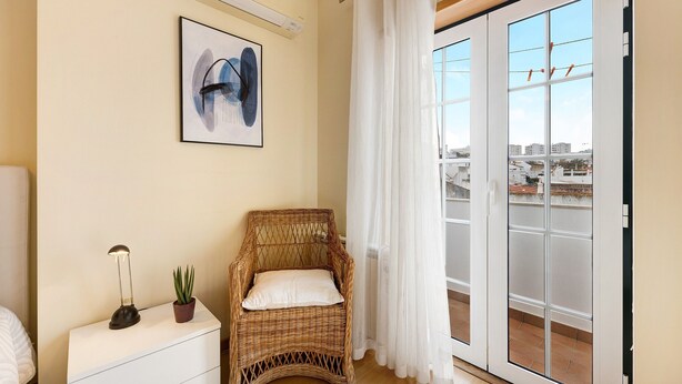 A room with a wicker chair, a white bedside table, a potted plant, a framed picture, and a sliding glass door leading to an outdoor balcony. - Holiday Apartment "Tourismer Centro" in City Center with Shared Terrace & Wi-Fi (Lagos)