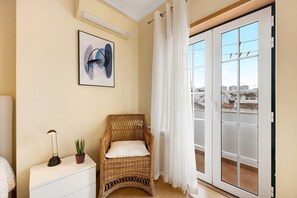 Una stanza con una sedia in vimini, un comodino bianco, una pianta in vaso, un quadro incorniciato e una porta scorrevole in vetro che dà su un balcone esterno.