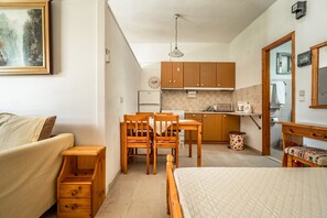 A compact room with a bed, a small wooden table, a chair, a kitchenette, and a painting on the wall.