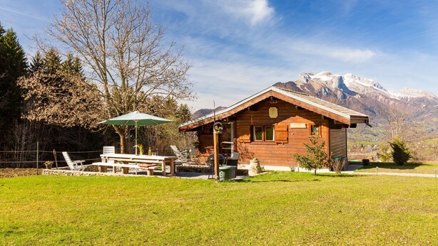 A wooden cabin with a patio, surrounded by a grassy area and trees, with mountains in the background. - Chalet "Sauvage" with Mountain Views, Private Terrace and Wi-Fi. (FAVERGES-SEYTHENEX)