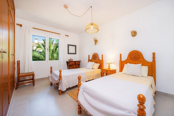 A bedroom with two wooden beds, a window with a view of greenery, a wooden chair, and a small table. - Chalet 'Casa Inés' with Private Terrace, Wi-Fi and Air Conditioning (Cala Murada)