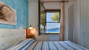 A bedroom with a bed, a nightstand, a lamp, a window with a view of the sea, and a wall with a painting.