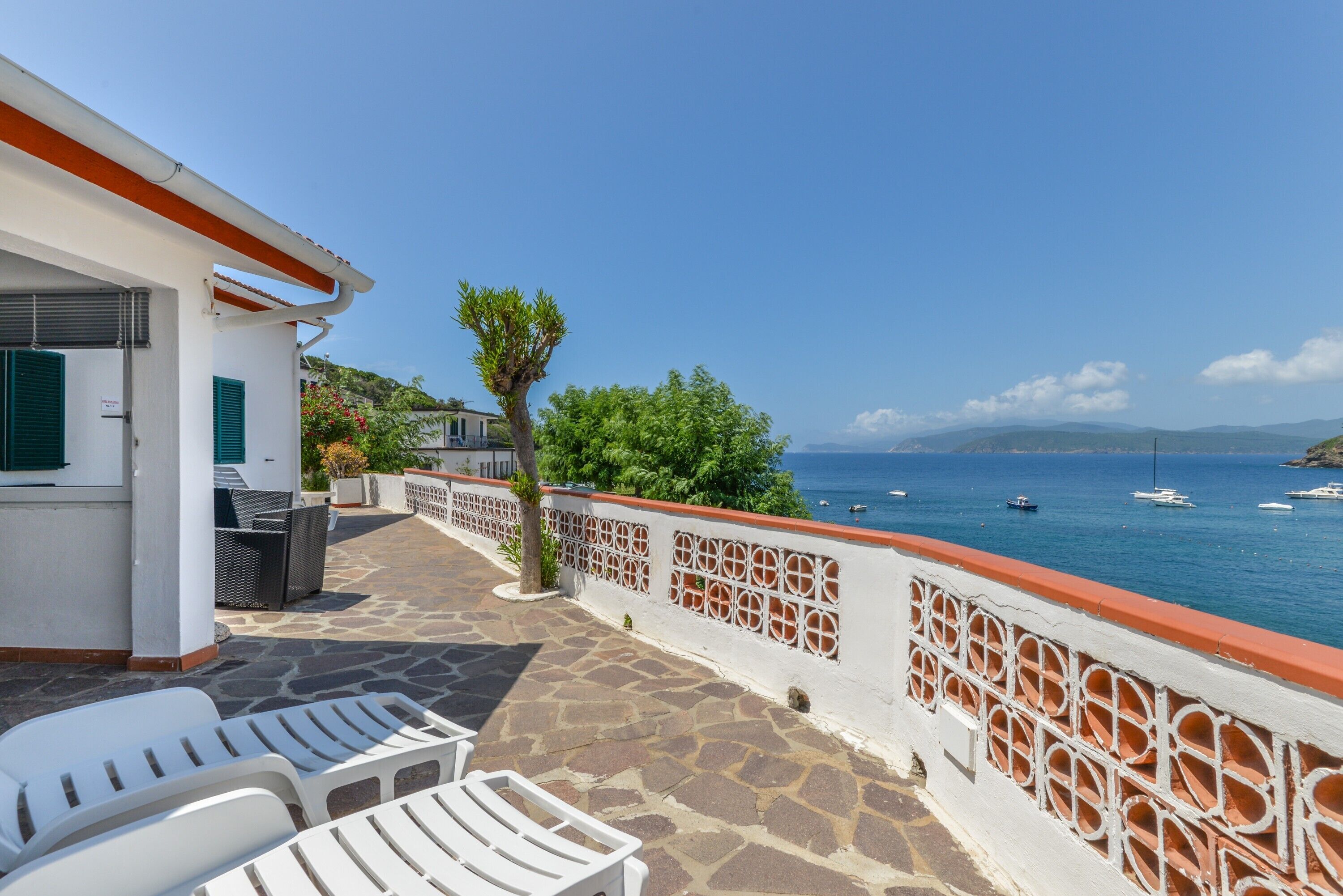 Eine Terrasse mit Steinboden, weißen Liegestühlen, einem kleinen Baum und Blick auf das Meer mit Booten.