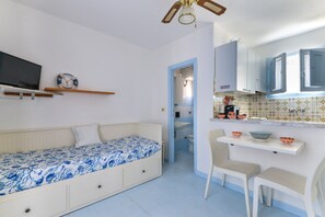 Habitación pequeña con cama, televisor, una kitchenette con pileta y alacenas, y un acceso a otra habitación.