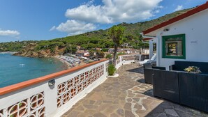 Un hôtel situé sur la côte, doté d’une terrasse pavée de pierres, d’un bâtiment blanc avec une porte verte et offrant une vue sur la plage et la mer.